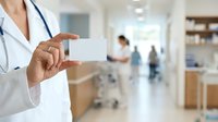 Doctor in white coat holding blank business card in medical clinic office environment with patients and staff in background.