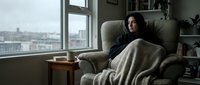 Young Asian woman relaxing in cozy armchair with blanket by window overlooking city skyline. Comfortable home interior for wellness and self-care concepts.