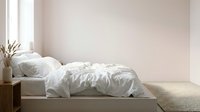 Unmade bed with white crumpled bedding, wooden nightstand with dried flowers in vase, minimalist bedroom interior with natural light from window.