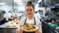 Female chef with blonde hair in professional kitchen holding colorful plated dish, restaurant staff working in background, culinary expertise and food service.