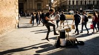 Street musician performing drums in urban plaza with pedestrians and historic architecture in background during daytime.