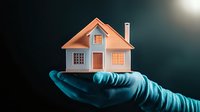Small residential house model held in glowing blue gloved hand against dark background, representing real estate, property protection, and home insurance concept.
