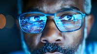 Close up of African American male programmer wearing glasses with reflection of programming code, blue digital technology glow illuminating focused eyes and face.