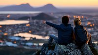 Young couple sitting on mountain peak overlooking city lights at sunset, sharing peaceful moment together while pointing at distant view, romantic outdoor adventure.