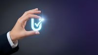 Hand holding glowing shield with checkmark icon representing cybersecurity protection and data safety for business technology concepts.