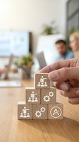 Hand placing wooden block with leadership icon on stacked pyramid of blocks featuring teamwork, gears, and management symbols. Office setting with blurred colleagues and computer in background.