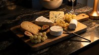 Assorted cheeses and crackers arranged on wooden board with grapes and knife on dark granite countertop for entertaining and appetizer preparation.
