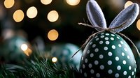 Green polka dot Easter egg with bunny ears decoration against blurred Christmas lights background, creating festive holiday atmosphere.