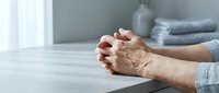 Senior woman hands clasped together in prayer or meditation pose on white table in peaceful home interior setting for wellness and spirituality concepts.