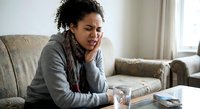 Young African American woman experiencing headache pain sitting on living room couch holding head in discomfort at home during daytime.