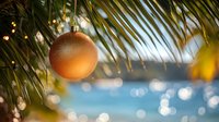 Golden Christmas ornament hanging from tropical palm fronds with blurred ocean and bokeh lights creating festive holiday vacation atmosphere.