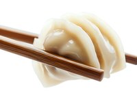 Asian dumplings held by wooden chopsticks on white background, close up view showing delicate folds and translucent wrapper texture of traditional dim sum.