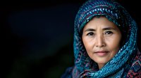 Mature Asian woman wearing traditional patterned blue hijab with intricate embroidery details smiles gently against dark background, capturing cultural heritage and dignity.
