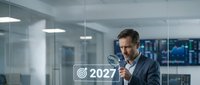Mature male professional in business attire holding digital target icon with 2027 text in modern office environment with glass partitions and city skyline view for corporate planning and future strategy concepts.