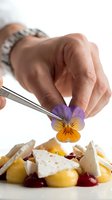 Adult male hand with tweezers placing purple and yellow pansy flower on gourmet plated dish with potatoes, cheese and berries for fine dining presentation.