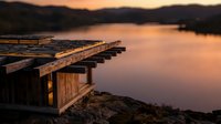 Wooden dock extending into calm lake at golden hour sunset, with warm orange and pink reflections on still water and distant forested shoreline.