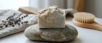 Handmade natural soap bar with exfoliating texture resting on smooth stone, surrounded by lavender, wooden brush and spa essentials for wellness and skincare routines.