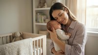 Young Caucasian mother tenderly holding sleeping newborn baby in bright nursery room with crib and natural window light for parenting content.