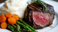 Juicy medium rare beef tenderloin steak served with creamy mashed potatoes, glazed baby carrots, green asparagus and fresh rosemary garnish on white plate.