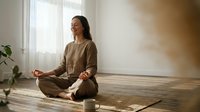 Young Asian woman practicing meditation in lotus position on yoga mat in bright modern home interior with natural sunlight streaming through window.