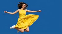 Young woman in bright yellow dress jumping with arms spread against clear blue sky expressing joy and freedom in summer outdoor lifestyle moment.