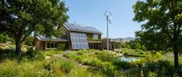 Modern eco-friendly house with solar panels and wind turbine surrounded by native landscaping and pond for sustainable living concepts.