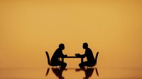 Two business professionals in silhouette having meeting discussion at table with chairs against warm orange gradient background for corporate presentations.