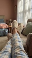 Cozy morning scene with legs in plaid pajamas and fuzzy slippers resting on armchair in warm bedroom with natural light streaming through curtains.