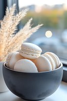 White macarons in dark ceramic bowl with pampas grass decoration against window background, soft natural lighting creates dreamy atmosphere for food photography.