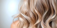 Close up view of wavy blonde highlighted hair with soft curls showing modern balayage technique and professional hairstyling on light background.