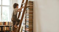 Young boy climbing wooden ladder to reach tall stack of books in bright library room for education and learning adventure.
