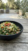 Tabbouleh salad with fresh parsley, tomatoes, and bulgur wheat served in black bowl on wooden table with olive trees and Mediterranean garden in soft sunlight background.