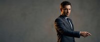 Confident businessman in dark suit and tie posing with crossed arms against textured gray background for corporate marketing materials.