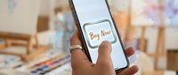 Hands holding smartphone with Buy Now button on screen, art studio background with watercolor splashes, mobile shopping and e-commerce concept.