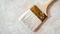 Wide paintbrush with white bristles and wooden handle on light textured background, close-up view showing brush detail and craftsmanship for home renovation.
