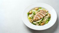 Grilled chicken breast sliced over Caesar salad with croutons and fresh greens served on white plate for restaurant menu or food blog.