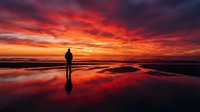 Silhouetted figure standing on wet beach at dramatic sunset with vibrant red and orange sky reflecting in water, conveying solitude and contemplation during golden hour.