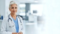 Confident mature female doctor with gray hair and stethoscope smiling in modern hospital corridor wearing white coat and blue scrubs for healthcare marketing.