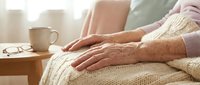 Elderly woman's hands resting on knit blanket in cozy home setting with warm morning light and coffee cup on wooden table nearby.