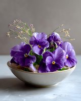 Purple pansies and delicate wildflowers arranged in ceramic bowl against neutral background, creating soft spring composition for floral and garden designs.