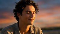 Young Hispanic man with curly hair smiling warmly during golden hour sunset, casual shirt, natural lighting for lifestyle and portrait photography.
