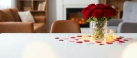 Red roses in crystal vase on white table with scattered petals in modern living room interior for romantic dinner date celebration.
