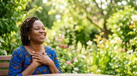 Woman with dark skin tone and dreadlocks wearing blue patterned clothing, sitting on wooden bench in lush green garden with sunlight filtering through foliage, expressing contentment and peace in nature.