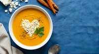 Creamy carrot soup with heart-shaped cream garnish and fresh herbs in white bowl on blue background with ingredients around for healthy comfort food.
