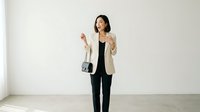 Young woman with dark hair, East Asian ethnicity, wearing a beige blazer over black clothing and holding a small black shoulder bag, standing against white wall background for business or fashion use.