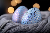 Speckled blue Easter eggs with golden dots nestled in cozy gray knitted fabric, soft bokeh lights creating magical holiday atmosphere in background.