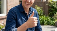 Smiling middle-aged Caucasian man in denim shirt giving thumbs up gesture outdoors near brick building with green foliage background for positive business content.