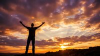 Silhouette of a man standing on rocky terrain with arms raised toward a dramatic golden sunset sky filled with glowing clouds.