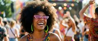 Young African American woman with afro hairstyle wearing sunglasses laughing joyfully at outdoor music festival with crowd dancing in background.