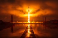 Dramatic rocket launch at sunset with fiery orange sky, creating golden reflection path on water as spacecraft ascends from coastal launch pad.